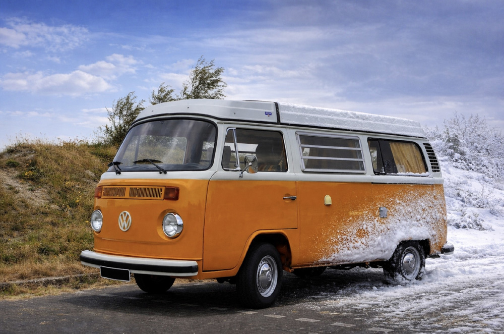 Orange and white vintage Volkswagen camper van parked on a road between a grassy hillside and a snowy landscape, showing a contrast between seasons