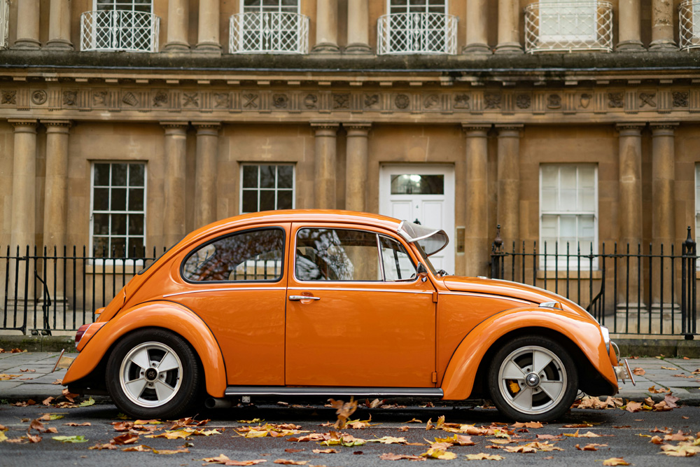 Orange vintage Volkswagen Beetle parked on a street in front of a neoclassical building with tall columns and autumn leaves on the ground