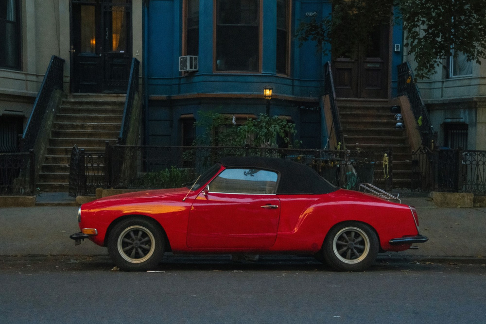 Red Karmann Ghia convertible parked on a residential street in front of brownstone buildings with stoops and a lit streetlamp