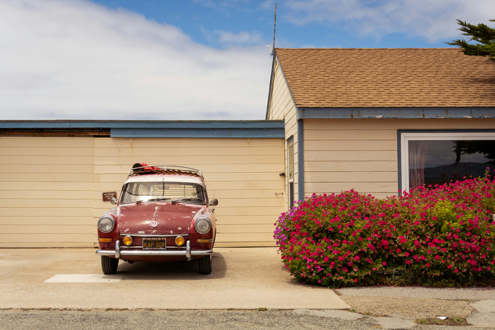 Red vintage Volkswagen Squareback parked in front of a wooden garage and a house with blooming pink flowers