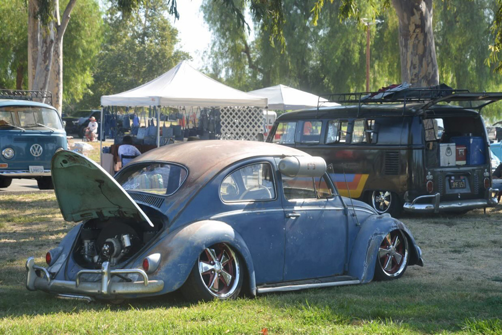 Weathered blue Volkswagen Beetle with open engine parked near two VW Buses and a vendor tent at the El Prado VW Show & Shine